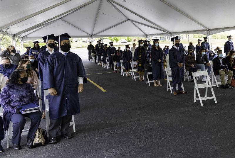 Students dressed in caps and gowns and wearing masks stand while audience members remain seated.