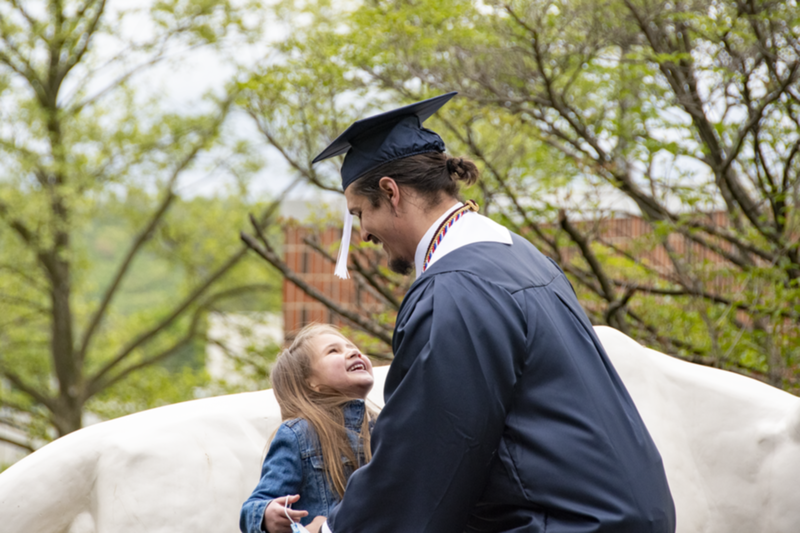 A man in academic regalia including picks up his small daughter in front of Schuylkill's lion shrine.